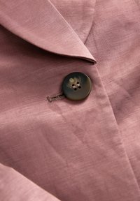 Nära bild av en rosa linneblazer med insnitt krage och en mörk, cirkulär knapp med fyra hål och ett marmorliknande mönster. Texturerad tygdetalj synlig.