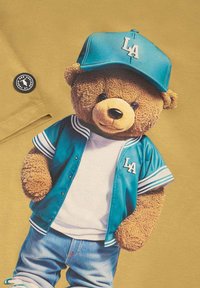 T-shirt graphique arborant un ours portant une casquette et une veste turquoise de LA, sur fond doré. Texture douce, design cartoon.
