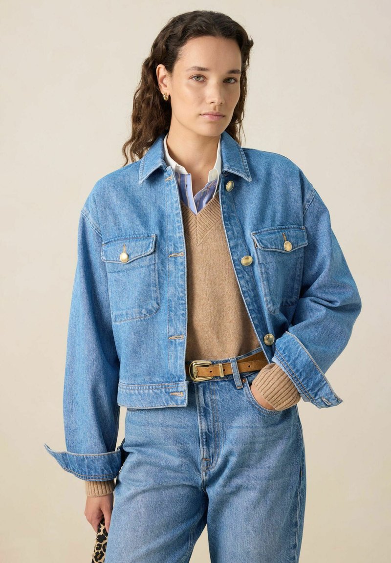 Lichtblauwe denim jacket met een cropped ontwerp, twee borstzakken, goudkleurige knopen en oversized mouwen, gelaagd over een tan-kleurige trui.