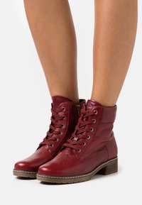 Botas de tobillo de cuero rojo con diseño de cordones, punta redonda y un tacón bajo apilado. Presentan ojales de metal y suela de goma cosida.