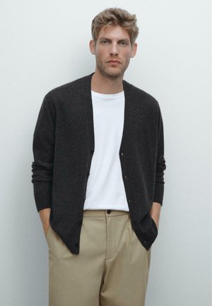Dark gray cardigan with a V-neckline, button closure, and long sleeves, worn over a white shirt and beige trousers. Soft texture.