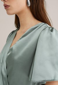 Green satin blouse with a V-neck and puff sleeves, featuring smooth texture and draped design elements. Silver geometric earrings visible.