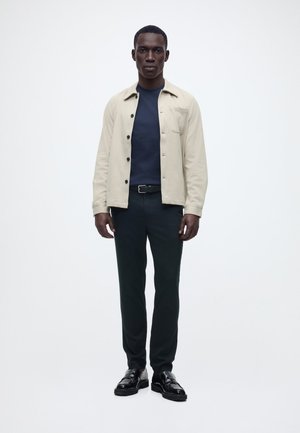 Mand står vendt fremad iført en beige knappet jakke over en marineblå skjorte, sorte bukser, sort bælte og sorte læder loafers.