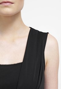 Nahaufnahme des unteren Gesichts einer Person, Hals, Schulter und oberer Brustbereich, die ein schwarzes ärmelloses Oberteil mit einer mit Spitze verzierten Halslinie trägt.