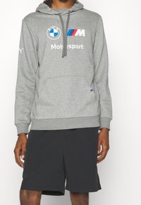 Sudadera gris con un bolsillo frontal, que presenta los logos en blanco de BMW y Motorsport, y una capucha con cordón. Combinada con pantalones cortos negros.