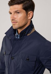 Giacca blu navy con colletto alto, due tasche sul petto con bottoni e dettagli marroni sul colletto. Realizzata in tessuto liscio e leggero.