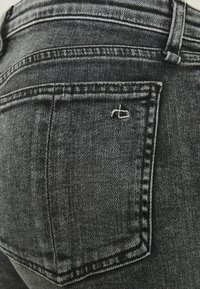 Jeans de mezclilla negra con un bolsillo trasero con costura blanca en contraste y un pequeño detalle de logo, que muestran un diseño y textura clásicos.