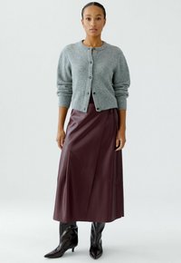 Vrouw staat gekleed in een grijze knopen cardigan, bordeaux leren midi-jurk en zwarte puntige kniehooglaarzen tegen een witte achtergrond.