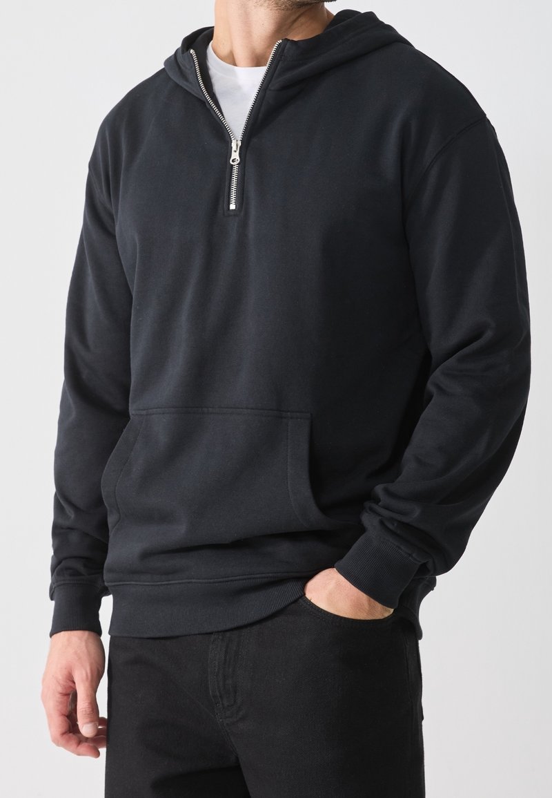 Sweat à capuche noir avec fermeture éclair d'un quart, poche ventrale, poignets côtelés, et une texture douce et lisse. Porté avec un jean foncé.