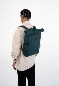 Matt zöld rolltop hátizsák, állítható pántokkal, elülső csattal, és oldalsó zsebekkel a kényelmes tárolás érdekében.