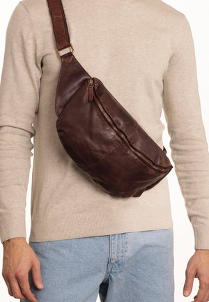 Brown leather sling bag with curved design, side zipper, and adjustable strap. Soft texture and casual style, worn over a light sweater.