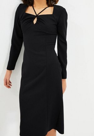 Czarna dopasowana sukienka z długimi rękawami, kwadratowym dekoltem, marszczonym przodem i wycięciem w kształcie klucza. Tkanina wydaje się gładka. Długość sukienki sięga poniżej kolan.