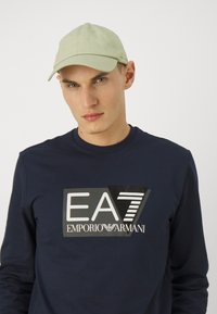 Maglietta a maniche lunghe blu navy con un grande logo brandizzato in bianco e nero, abbinata a un cappellino verde chiaro. Materiale in cotone, design casual, forma aderente.