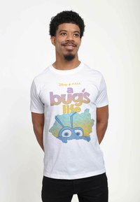 Camiseta de algodón blanca con un personaje de dibujos animados azul y ojos grandes, rodeado de follaje verde y amarillo, y el texto "la vida de un bicho".
