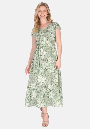 Nő zöld-fehér mintás, rövid ujjú maxi ruhában, derekán megkötővel, állva és mosolyogva, egyszínű háttér előtt.