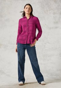Camicia fucsia con bottoni e motivi geometrici neri, abbinata a jeans larghi blu scuro e sneaker chiare su uno sfondo testurizzato.