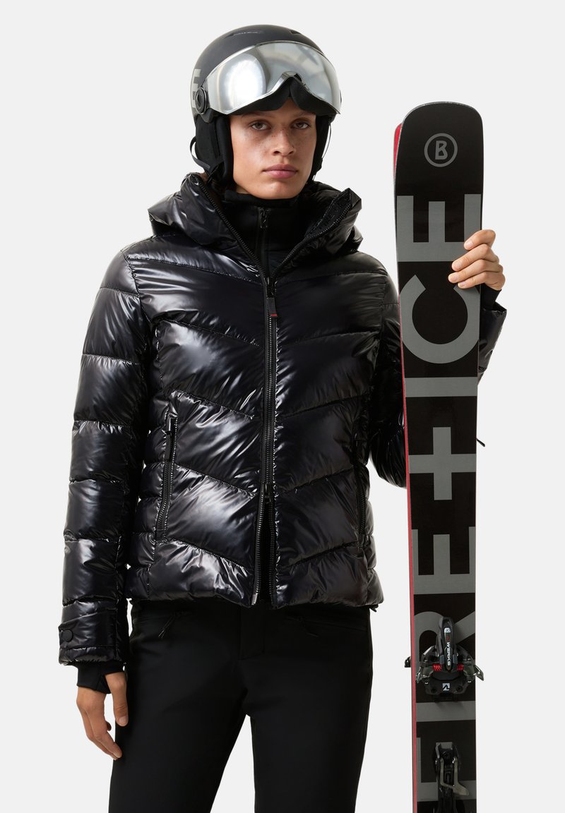 Zwarte pufferjas met een glanzende afwerking, chevronpatroon, ritssluiting aan de voorkant en capuchon. Model houdt een zwarte ski vast met grijze letters, draagt een helm en een skibril.