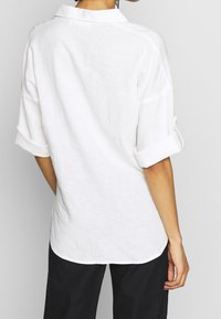 Camicia di lino bianco con maniche arrotolate e colletto; orlo leggermente curvo; texture leggera. Focus sulla vista posteriore e dettagli delle maniche.