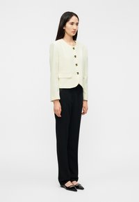 Εκρού σακάκι με στρογγυλή λαιμόκοψη, πέντε διακοσμητικά κουμπιά και δύο μπροστινές τσέπες, συνδυασμένο με μαύρο παντελόνι και επίπεδα μυτερά παπούτσια.