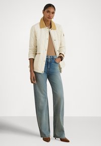 Geacă matlasată de culoare bej deschis, cu guler muștar, purtată peste un top căprui și blugi albastri cu picioare largi. Pantofii maro cu toc completează ținuta.