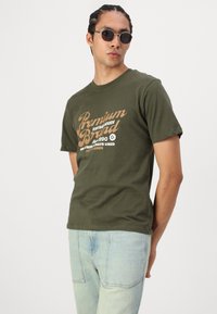 Olivno zelena bombažna majica s kratkimi rokavi, okrašena s svetlo rjavim grafičnim tiskom. Ujemajoči lahki modri jeans.