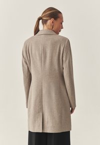 Pikk beež villane mantel klassikalise kraega, sirge lõikega ja tagumise tuuleaugu detailiga, nähtud tagant. Tekstuuriga materjal, minimalismi stiilis disain.