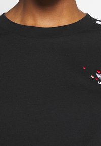 Närbild av en person som bär en svart t-shirt med rund halsringning, försedda med ett litet rött hjärta och vit grafisk brodering på bröstet.