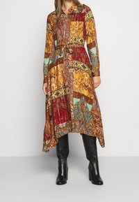 Vrouw in een jurk met lange mouwen, asymmetrische zoom, met kleurrijke paisley patchworkprint en zwarte, kniehoge, hakkenlaarzen.