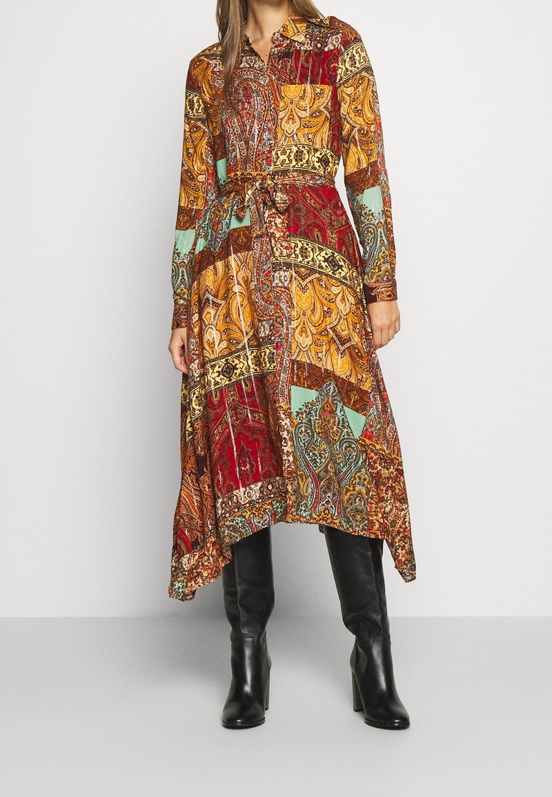Vrouw in een jurk met lange mouwen, asymmetrische zoom, met kleurrijke paisley patchworkprint en zwarte, kniehoge, hakkenlaarzen.