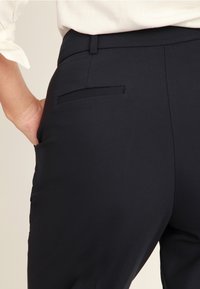 Person som bär mörka skräddarsydda byxor och en vit skjorta med ena handen i bakfickan, sedd från midjan och neråt.