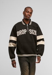 Zwart pullover sweatshirt met ribgebreide kraag, ritsluiting en de tekst "DROP SIZE". Beschikt over crèmekleurige strepen op de mouwen en de tailleband.