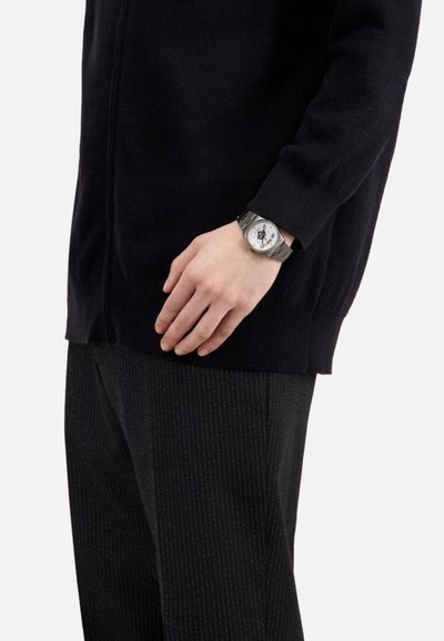 Reloj plateado con una esfera blanca redonda, acentos negros y una correa de metal, que se lleva en una mano que descansa sobre un suéter de punto oscuro y texturizado y pantalones a rayas.