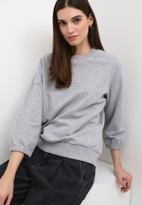 Felpa grigia chiaro con scollatura rotonda e maniche a sbuffo a tre quarti. Presenta polsini e orlo a coste elasticizzati. Indossata con pantaloni di jeans scuri.