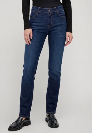 Personne portant un jean droit bleu foncé, un haut noir à manches longues et des chaussures en cuir noires sans lacets sur un fond uni.