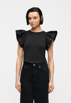 Γυναίκα με κοντά σκούρα μαλλιά που φοράει μαύρο κοντό τοπ με μεγάλα βολάν στα μανίκια στολισμένα με μικρές ασημένιες καρφίτσες, συνδυασμένο με μαύρο παντελόνι.