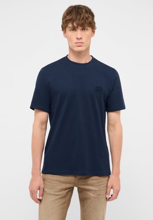 Junger Mann mit hellbraunen lockigen Haaren trägt ein schlichtes dunkelblaues T-Shirt mit kurzen Ärmeln und hellbraune Hose, steht vor weißem Hintergrund.