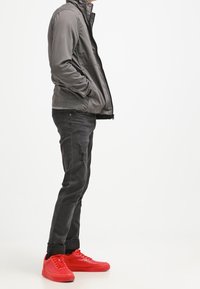 Szara kurtka na zamek z wysokim kołnierzem, czarne zniszczone dżinsy oraz jasnoczerwone trampki z teksturowanymi dodatkami. Widok z boku.