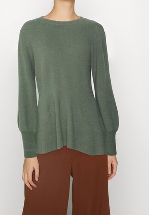 Femme portant un pull vert côtelé à manches longues avec un col rond et un pantalon large marron, coupé à la moitié du torse et à la moitié de la cuisse.