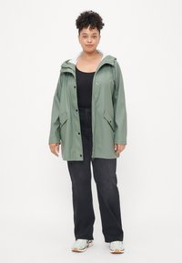 Πράσινο αδιάβροχο μπουφάν με κουκούλα, μπροστινό φερμουάρ και κουμπιά. Φοριέται πάνω από μαύρο τοπ, συνδυασμένο με μαύρο παντελόνι και ανοιχτόχρωμα αθλητικά παπούτσια.