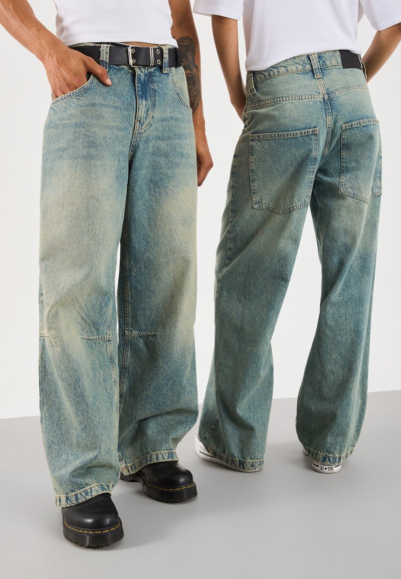 Vaqueros anchos de pierna ancha en azul claro con acabado desgastado, bolsillos delanteros y traseros, y costuras visibles. Combinados con calzado en blanco y negro.