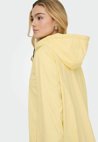 Rumena jopa z kapuco, sproščenega kroja in gladke teksture, z bočnimi žepi in črnimi kontrastnimi zadrgami.