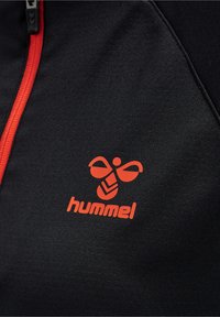 Chaqueta atlética negra con una cremallera roja, que presenta un destacado logo naranja de "hummel" y un diseño de mariposa en la zona del pecho izquierdo.