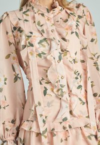Abito in chiffon floreale di un delicato rosa con rifiniture voluminose e chiusura con bottoni. Presenta un colletto alto, maniche lunghe e delicati motivi floreali.