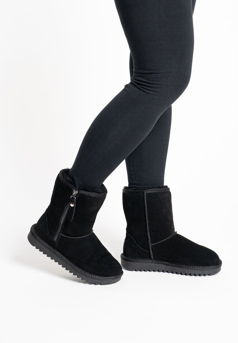 Personne portant des bottines noires à fermeture éclair latérale et des leggings noirs, debout avec un pied légèrement levé.