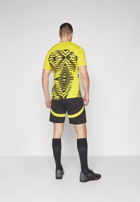 Gul atletisk tröja med ett svart geometriskt mönster på ryggen, kombinerad med svarta shorts med gula detaljer och svarta kompressionsstrumpor.