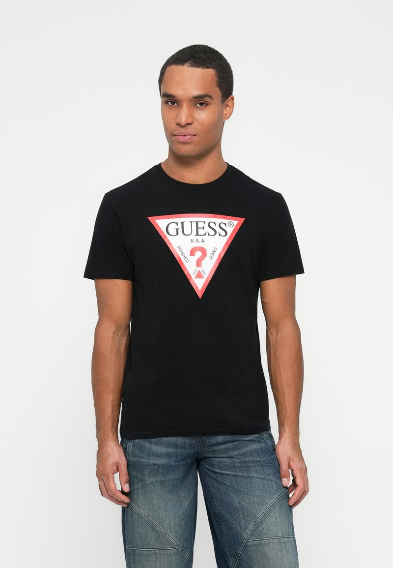 Tricou negru din bumbac, cu un logo triunghiular roșu mare și inscripția "GUESS U.S.A." și un semn de întrebare alb la centru. Croi lejer.