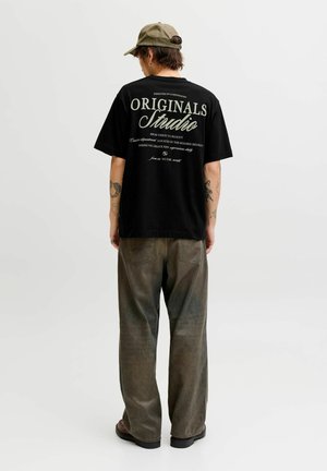 Svart t-shirt i bomull med vitt och grönt grafiskt tryck på baksidan, kombinerad med löst sittande, blekta olivgröna byxor. Bruna skor.