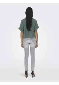 Losse groene blouse met opgerolde mouwen, gecombineerd met strakke lichtgrijze jeans en zwarte hoge hakken. Haar valt voorbij de schouders.