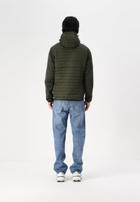 Olivgrüne, gesteppte Jacke mit Kapuze, getragen über hellblauen Jeans. Sie hat eine glatte Textur mit dezenten Nähdetails.