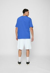 Blå bomull T-skjorte med korte ermer, kombinert med hvite shorts. Modellen står med ryggen til, iført svarte slip-on sko og hvite sokker.